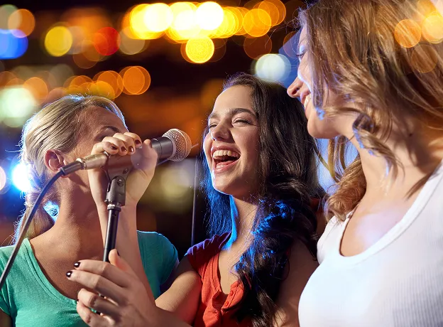 Trois jeunes femmes chantant au karaoké, riant, sur fond flou de lumières.